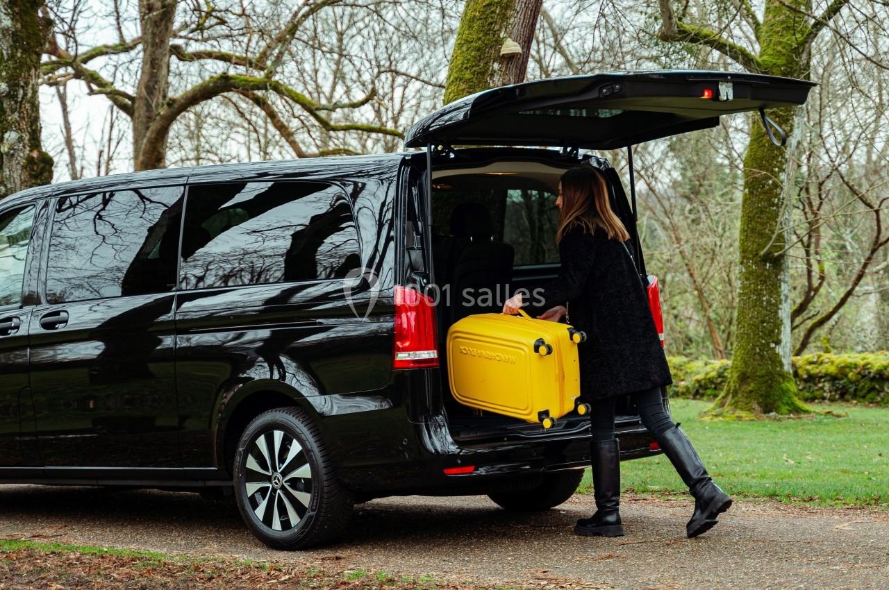 Une femme charge une valise jaune dans le coffre ouvert d'un van noir stationné dans un environnement boisé.
