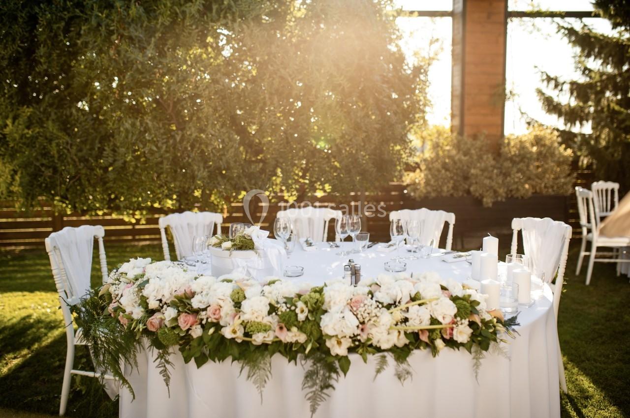 Table ronde décorée de fleurs blanches et roses, dressée pour un événement en plein air sous un arbre.
