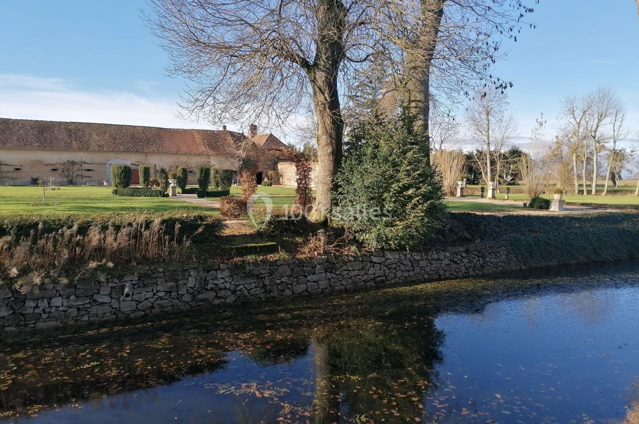 Vue d'un bâtiment en pierre entouré d'arbres, avec un petit cours d'eau au premier plan sous un ciel dégagé.