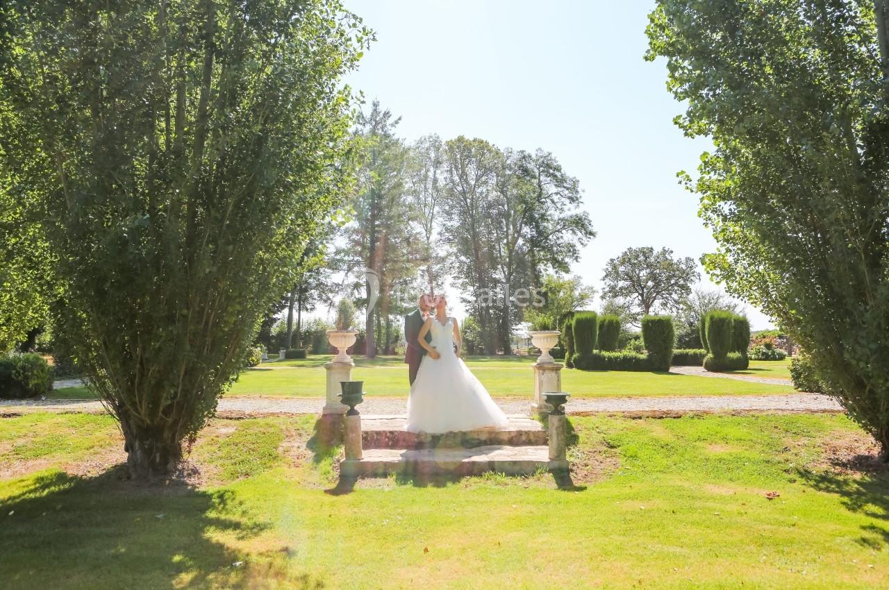 Un couple en tenue de mariage pose dans un jardin verdoyant, encadré par des arbres et baigné de lumière naturelle.