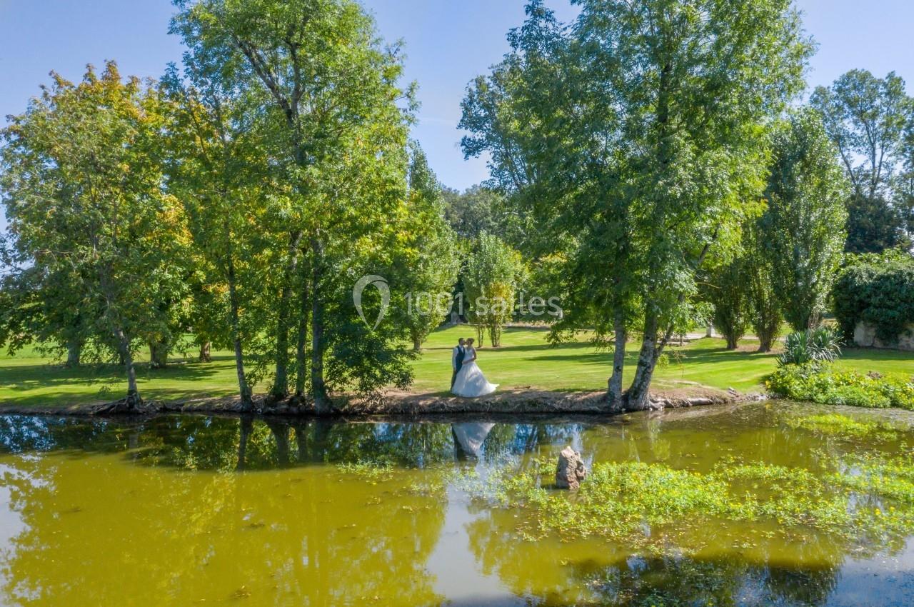 Un couple en tenue de mariage se tient sur une berge entourée d'arbres, près d'un étang calme.
