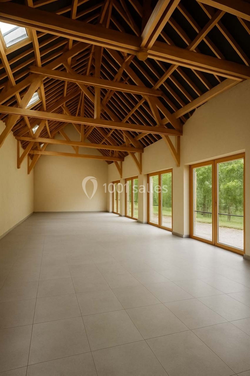 Grande salle vide avec un plafond en bois apparents, des murs clairs et de grandes fenêtres donnant sur un espace vert.