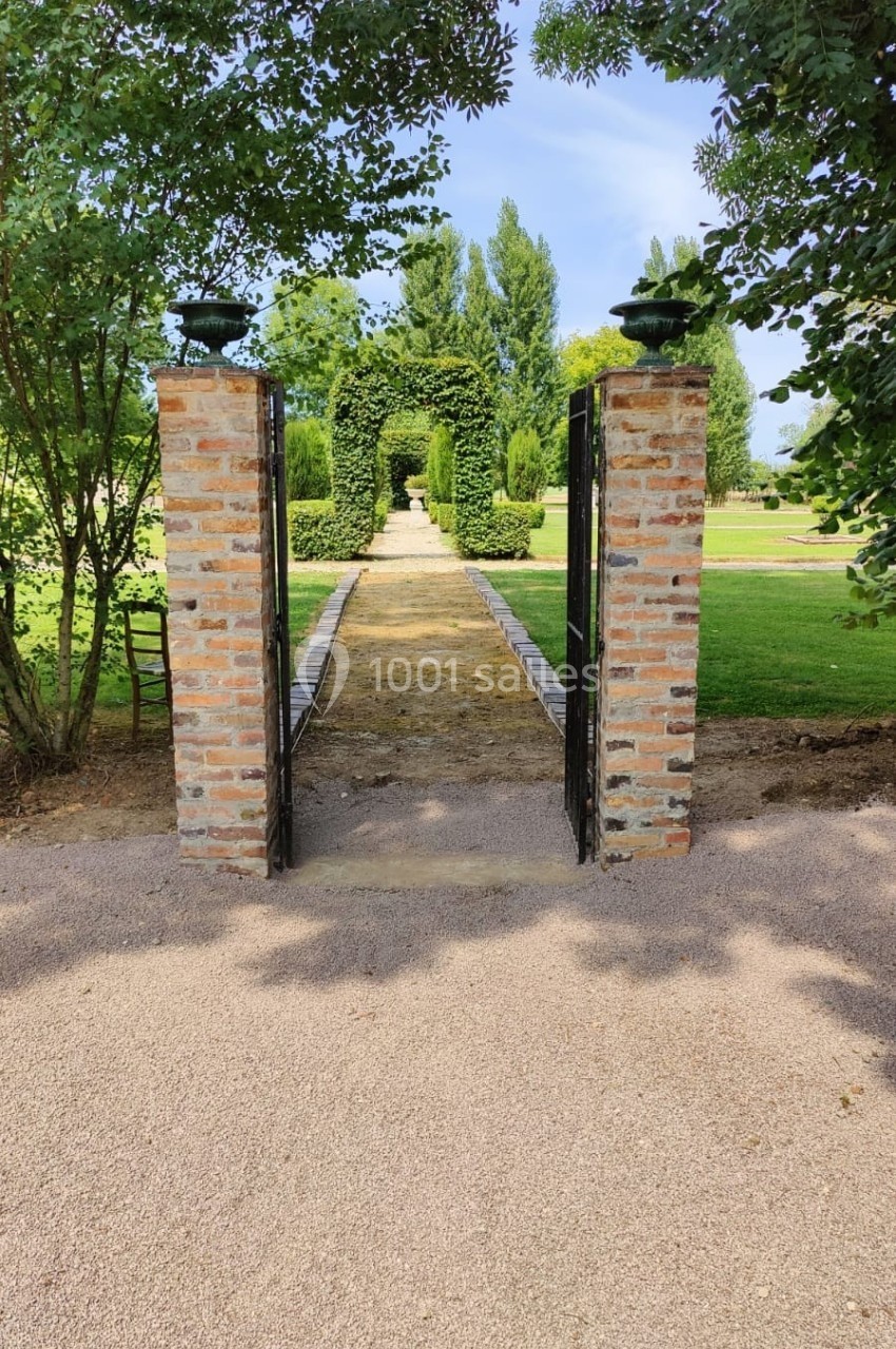 Allée bordée de pelouse et d'arbres, vue à travers une porte en briques et fer forgé dans un jardin.
