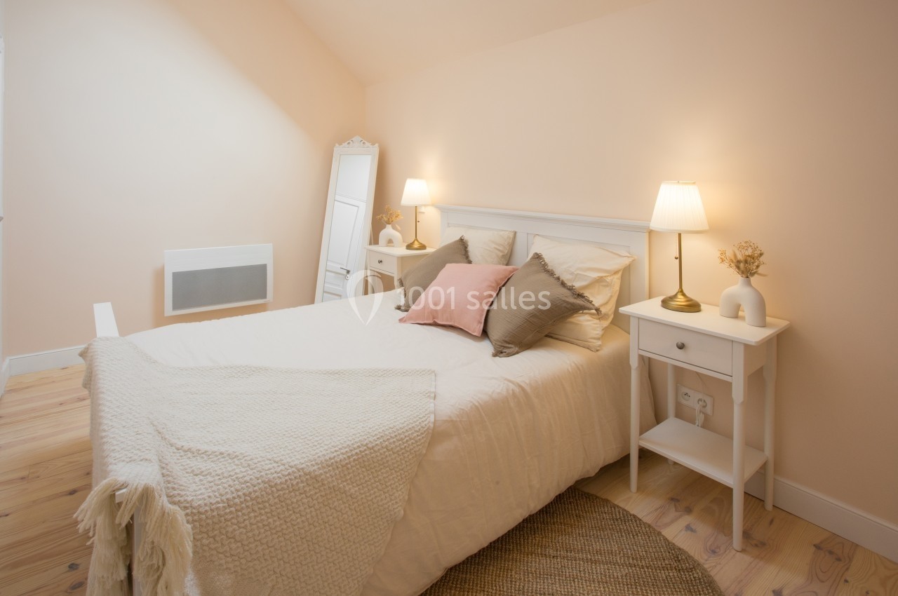 Chambre lumineuse avec lit double, coussins, tables de chevet blanches, lampes allumées et miroir sur pied.