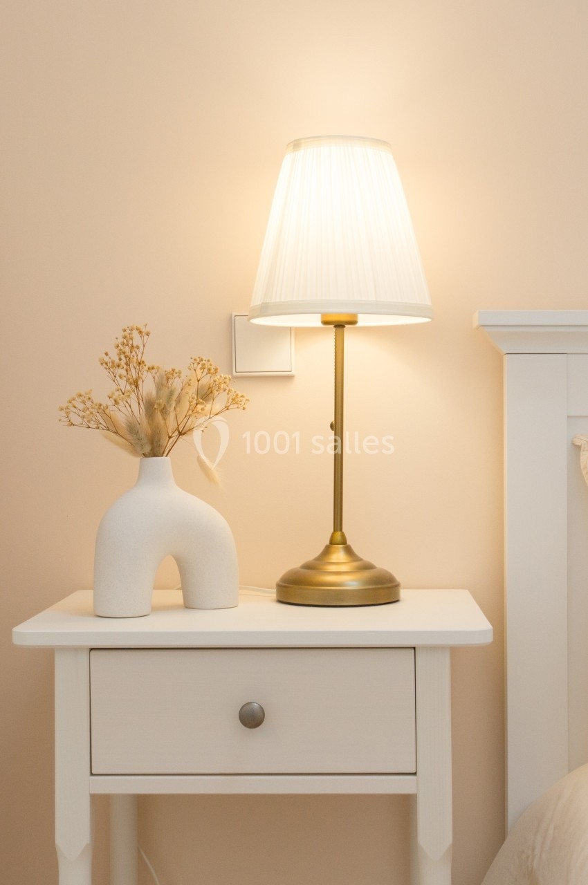 Lampe de chevet allumée sur une table blanche avec un vase décoratif contenant des fleurs séchées.