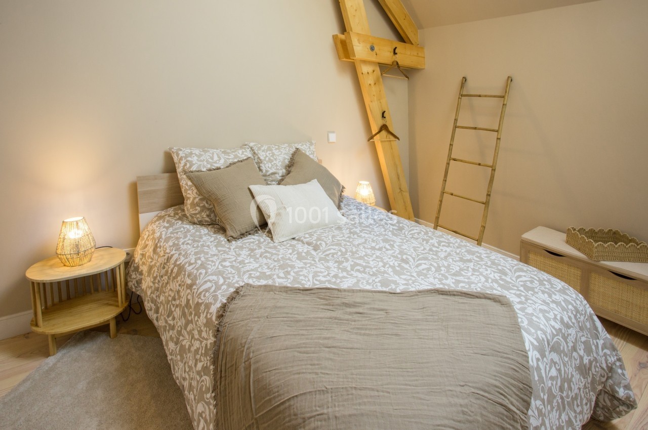Chambre avec lit double, linge beige et blanc, tables de chevet en bois, échelle décorative et lumière douce.
