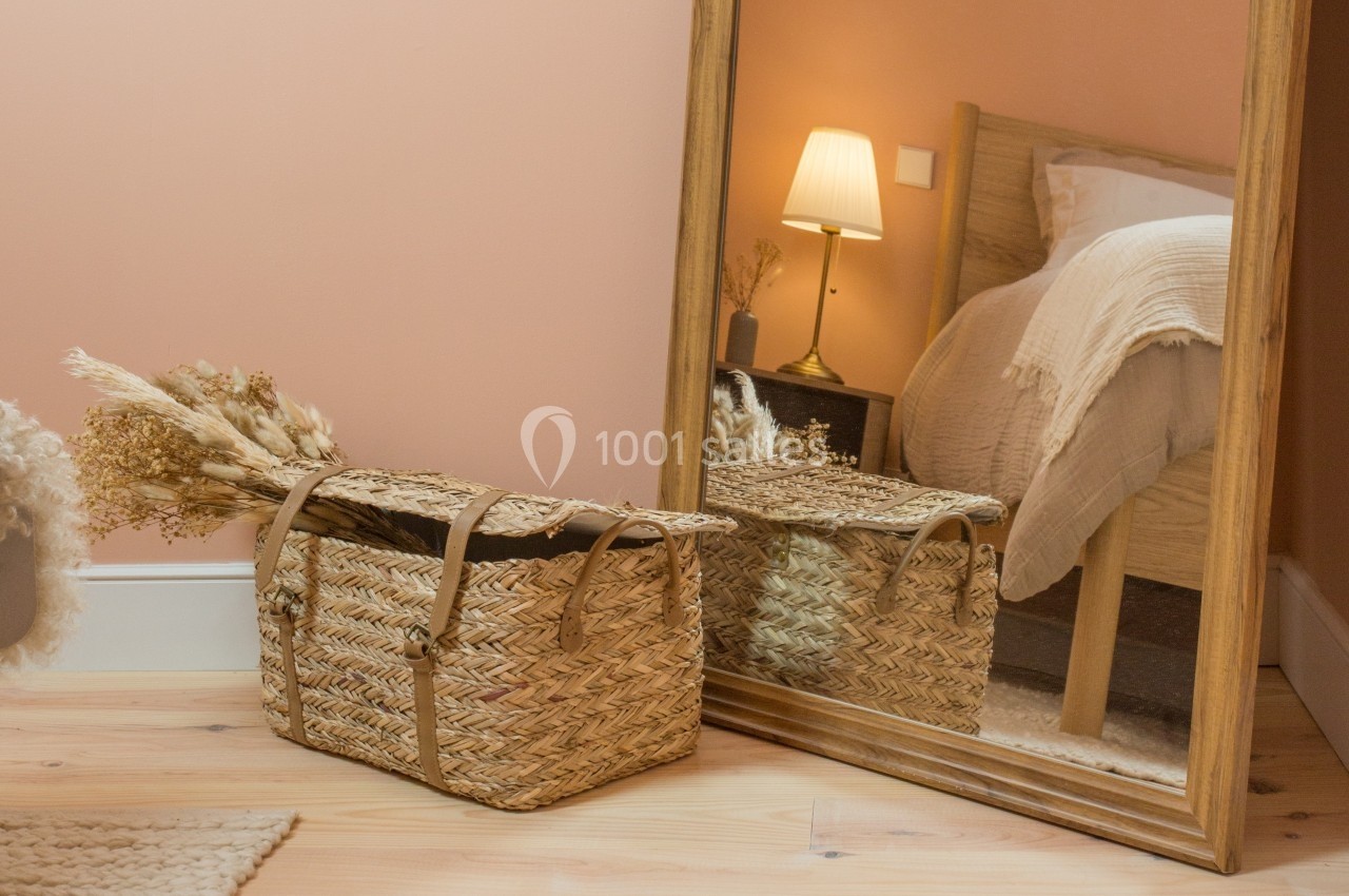 Panier en osier posé au sol devant un grand miroir, avec un lit et une lampe en arrière-plan dans une chambre.
