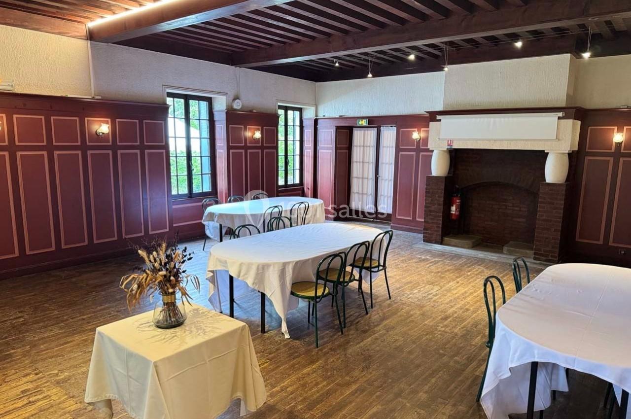Salle lumineuse avec parquet, murs en bois rouge, cheminée, tables dressées avec nappes blanches et chaises.