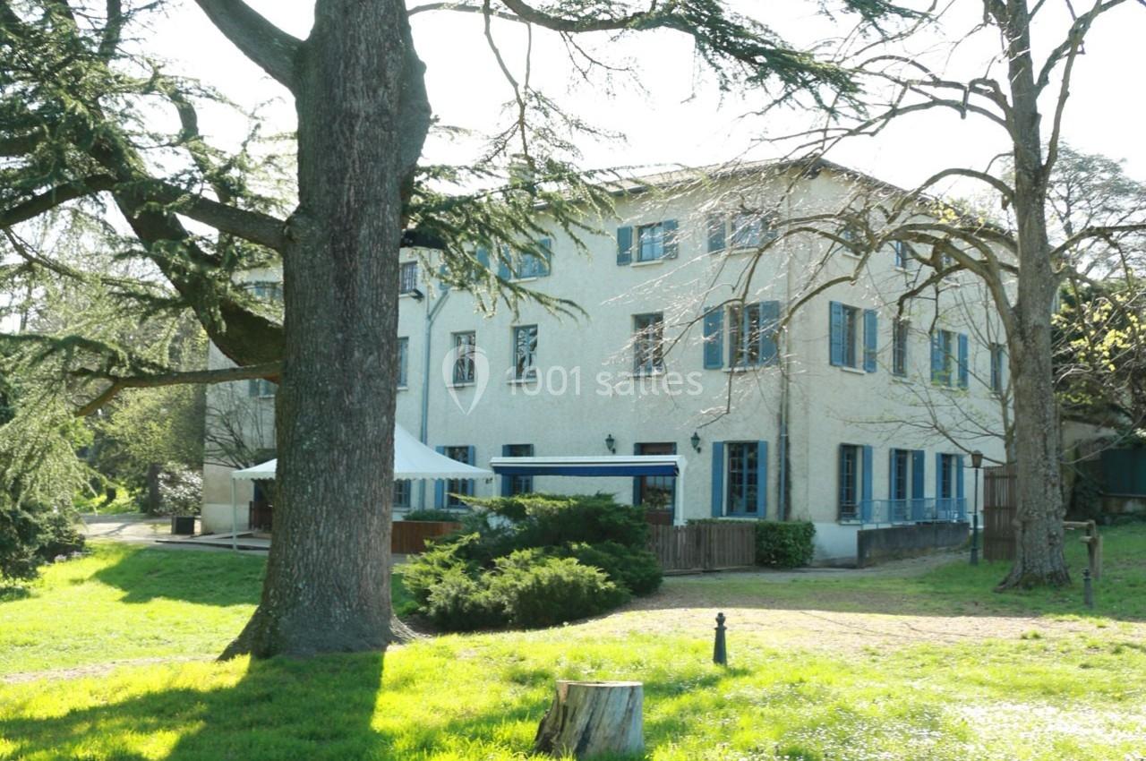 Grande maison blanche à volets bleus entourée d'arbres et d'une pelouse verdoyante sous un ciel clair.