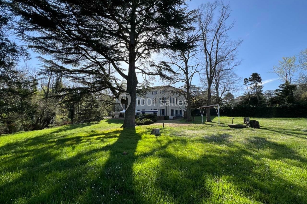 Grande maison blanche entourée d'arbres et d'une pelouse verdoyante sous un ciel bleu clair.