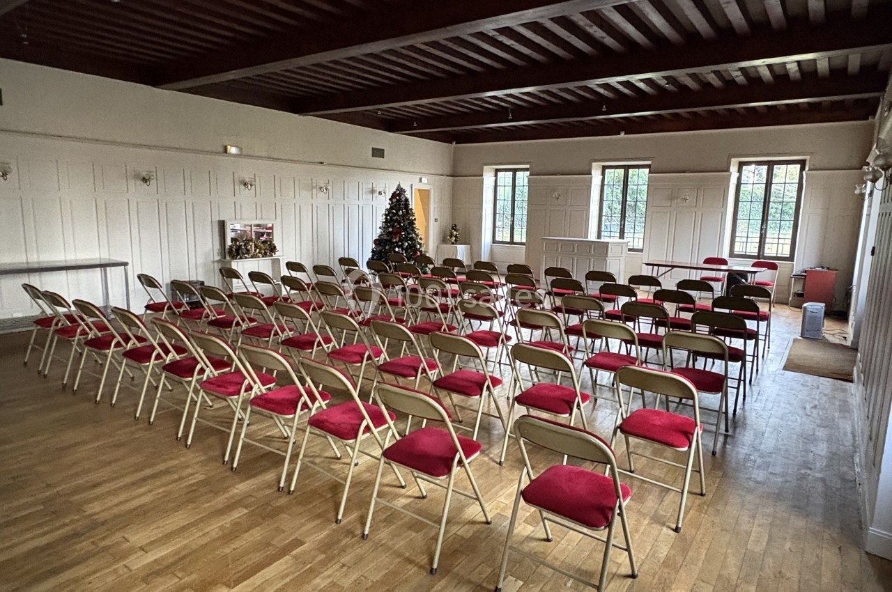 Salle avec parquet, rangées de chaises pliantes à assise rouge, sapin de Noël décoré et fenêtres laissant entrer la lumière.