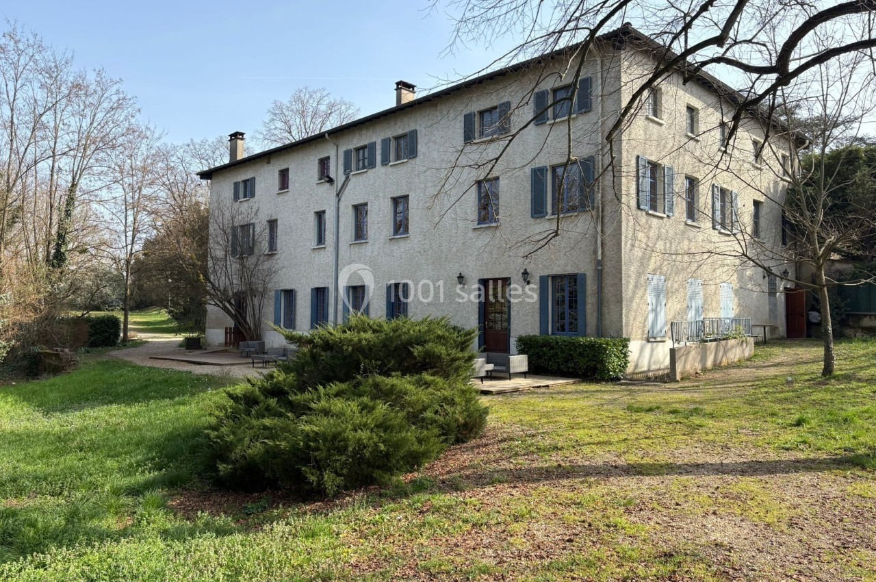 Grande maison blanche à deux étages avec volets bleus, entourée d'arbres et d'un jardin verdoyant.