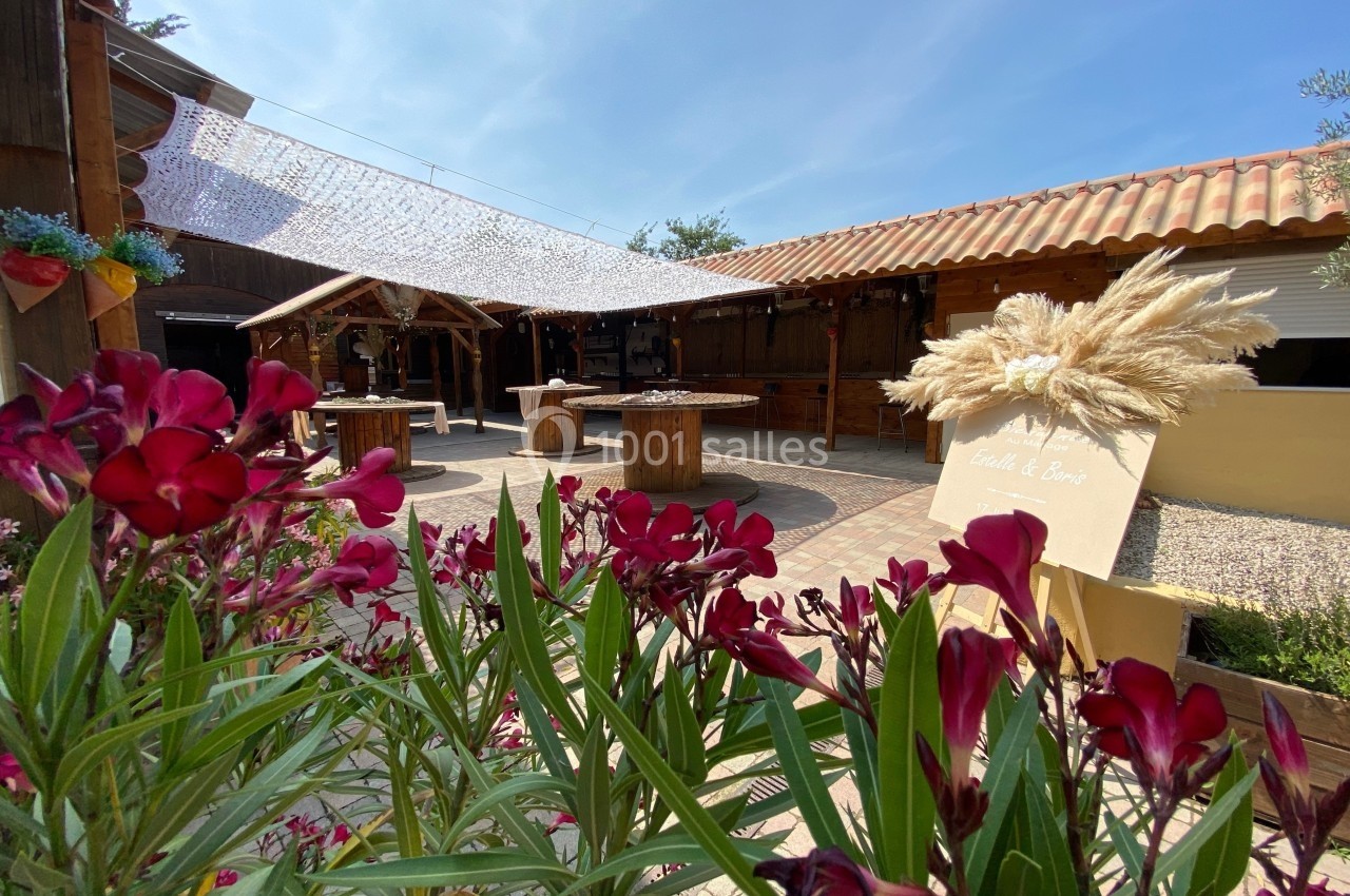 Espace extérieur avec tables en bois, fleurs au premier plan et voile d'ombrage sous un ciel dégagé.