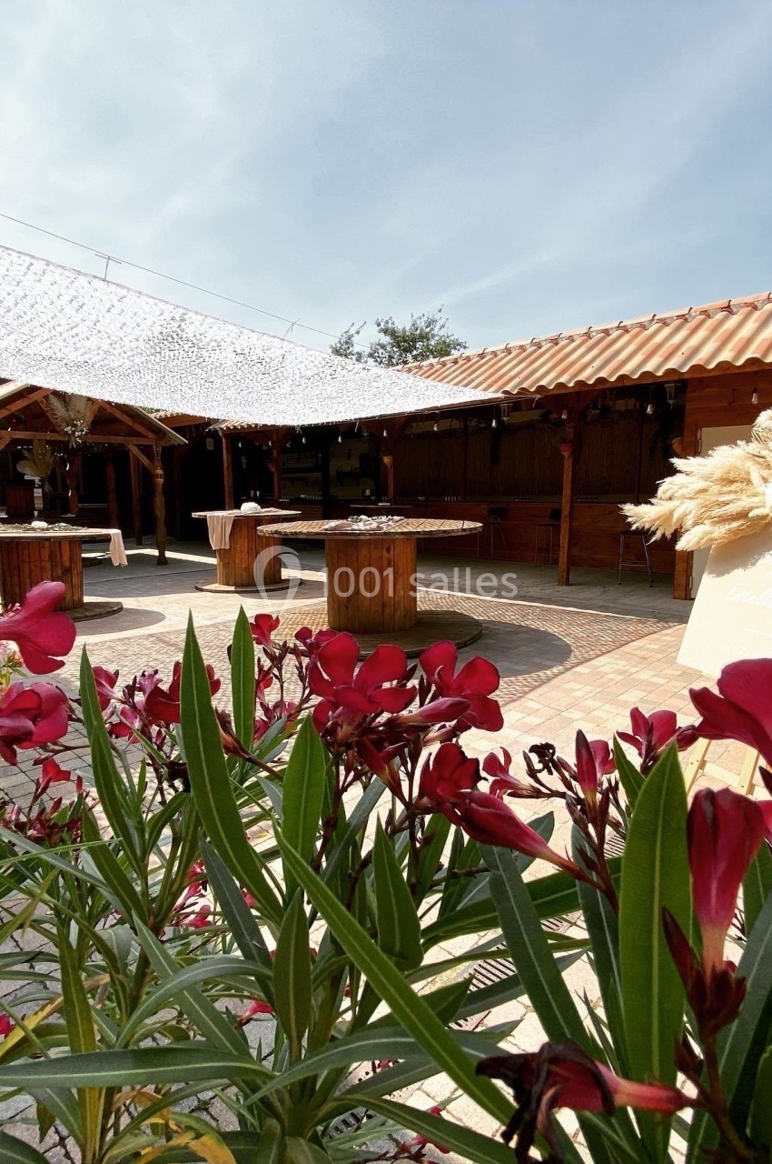 Cour extérieure ensoleillée avec tables en bois, décorations florales et voile d'ombrage, entourée de bâtiments en bois.