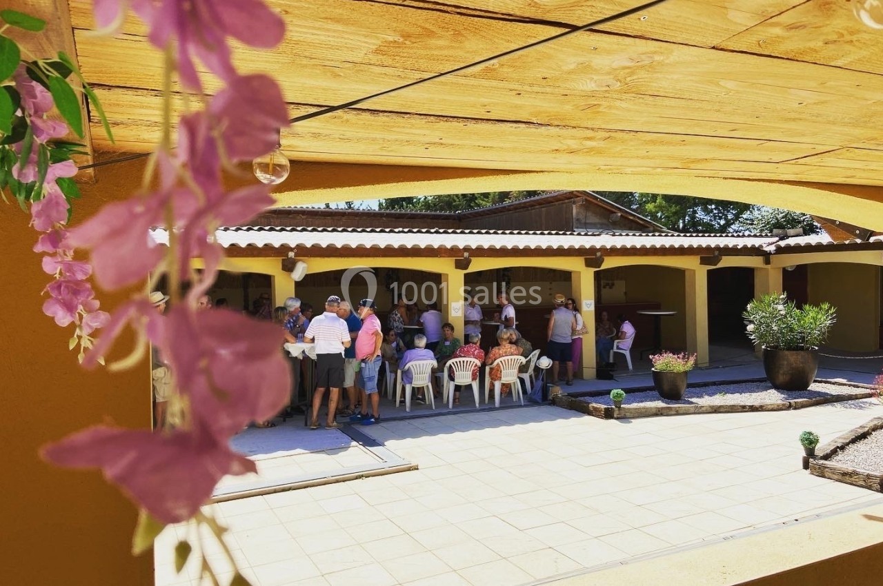 Un groupe de personnes assises et debout sous un auvent, dans une cour ensoleillée avec des fleurs au premier plan.