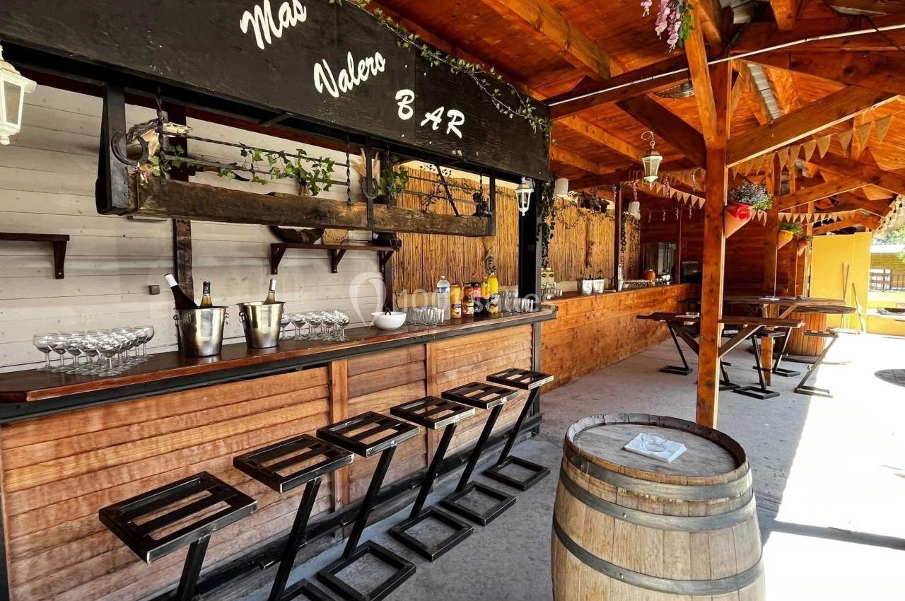 Bar en bois avec tabourets, tonneau en premier plan, étagères décorées et bouteilles exposées sous un toit en bois.