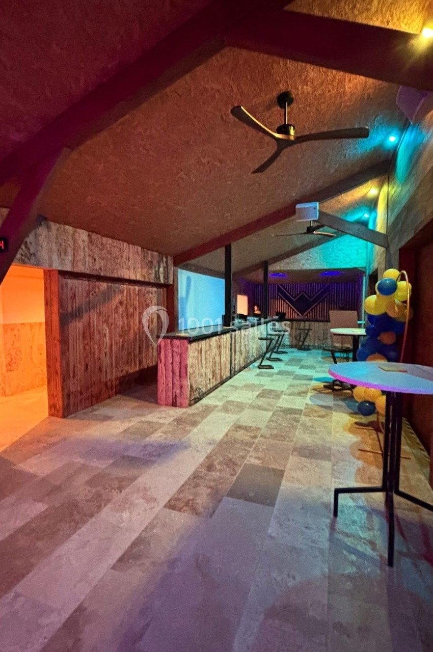 Salle avec un bar en bois, un plafond incliné, des éclairages colorés et des tables hautes.