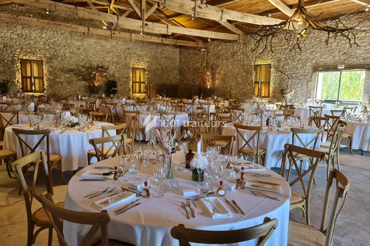 Salle de réception en pierre avec tables rondes dressées, nappes blanches et chaises en bois, éclairage chaleureux.