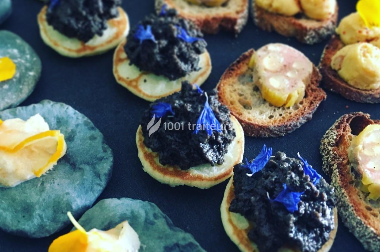 Assortiment de canapés garnis de tapenade, fleurs comestibles et autres ingrédients sur une table sombre.