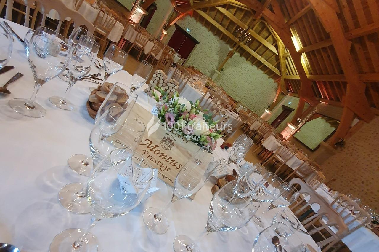 Salle de réception avec tables dressées, nappes blanches, verres, couverts et centre de table floral sous une charpente en…