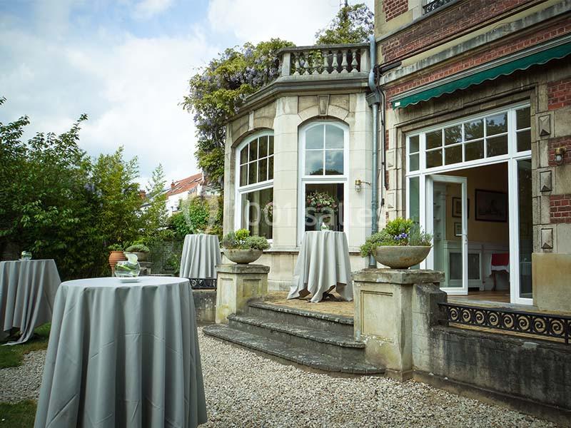 Terrasse d'une maison ancienne avec des tables hautes drapées de nappes grises, entourée de verdure.