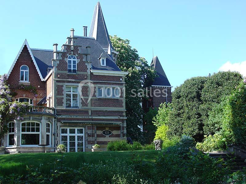 Grande maison en briques rouges avec des toits en ardoise, entourée d'arbres et d'un jardin verdoyant.