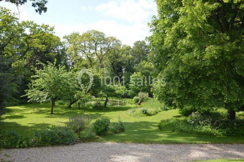 Jardin verdoyant avec pelouse, arbres feuillus et petit étang sous un ciel partiellement nuageux.