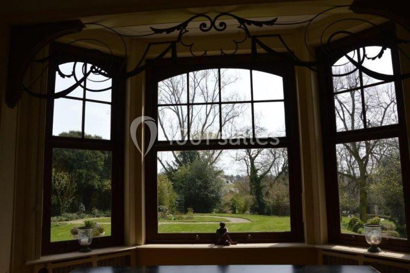 Vue depuis une fenêtre en baie donnant sur un jardin arboré avec pelouse et arbres dénudés en arrière-plan.