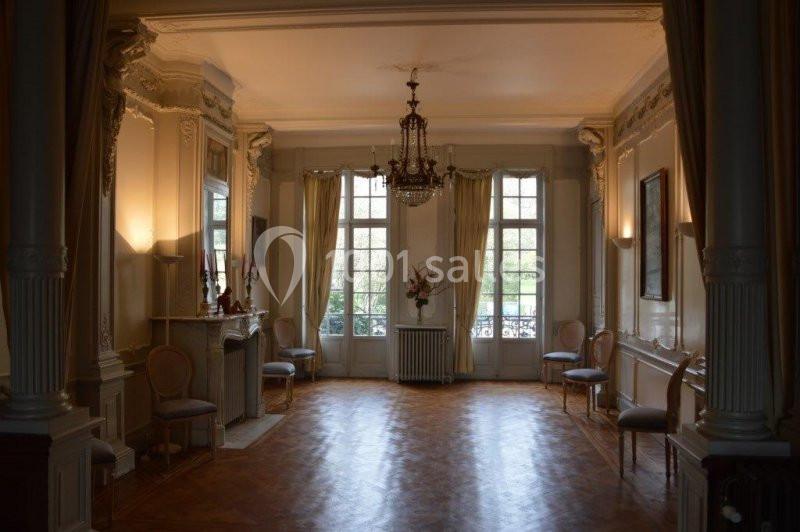 Salle élégante avec parquet, moulures, lustre central, grandes fenêtres et chaises disposées le long des murs.
