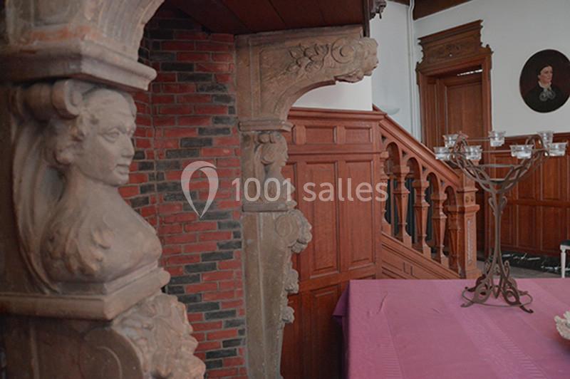 Détail d'une cheminée en pierre sculptée dans une pièce avec boiseries, escalier en bois et chandelier sur une table.
