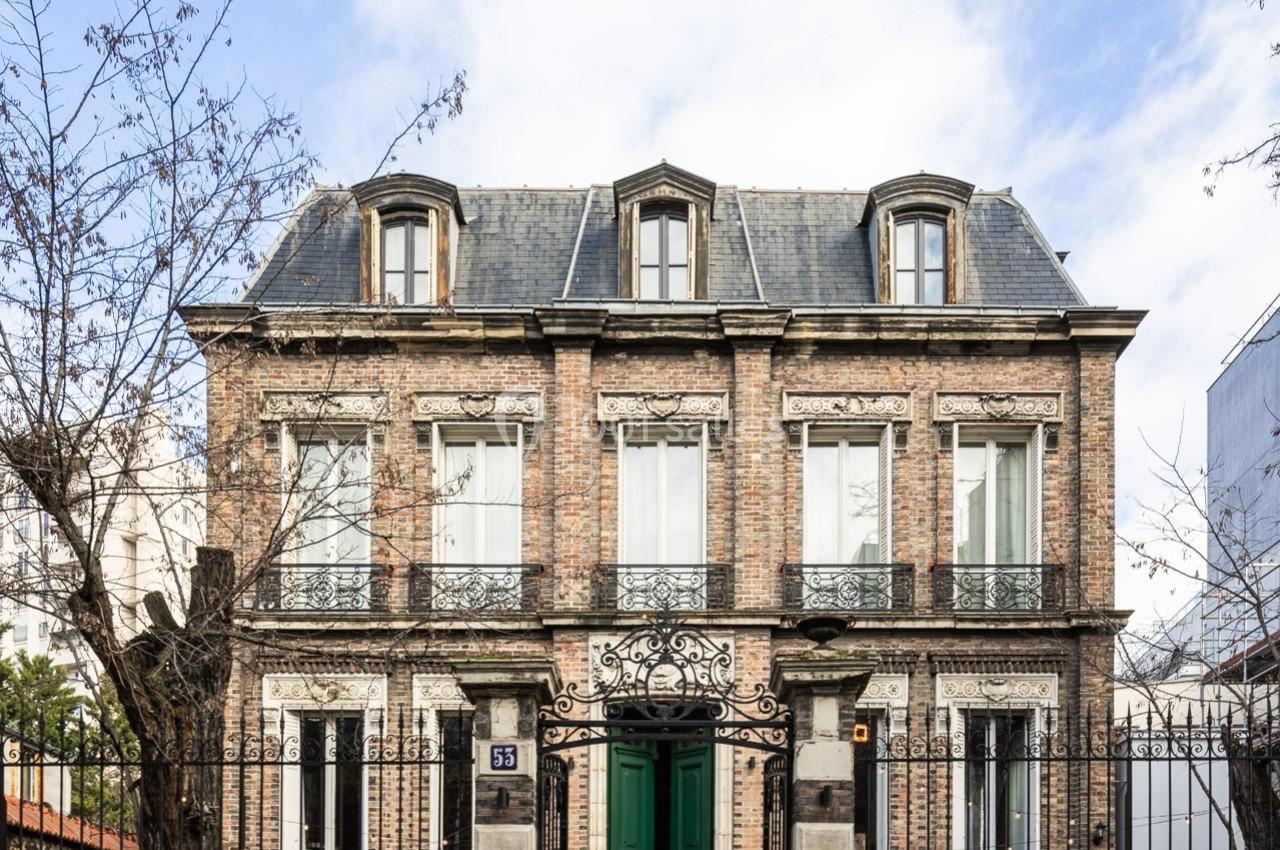 Façade d'une maison en briques à deux étages avec toit mansardé, portail en fer forgé et arbres dénudés devant.