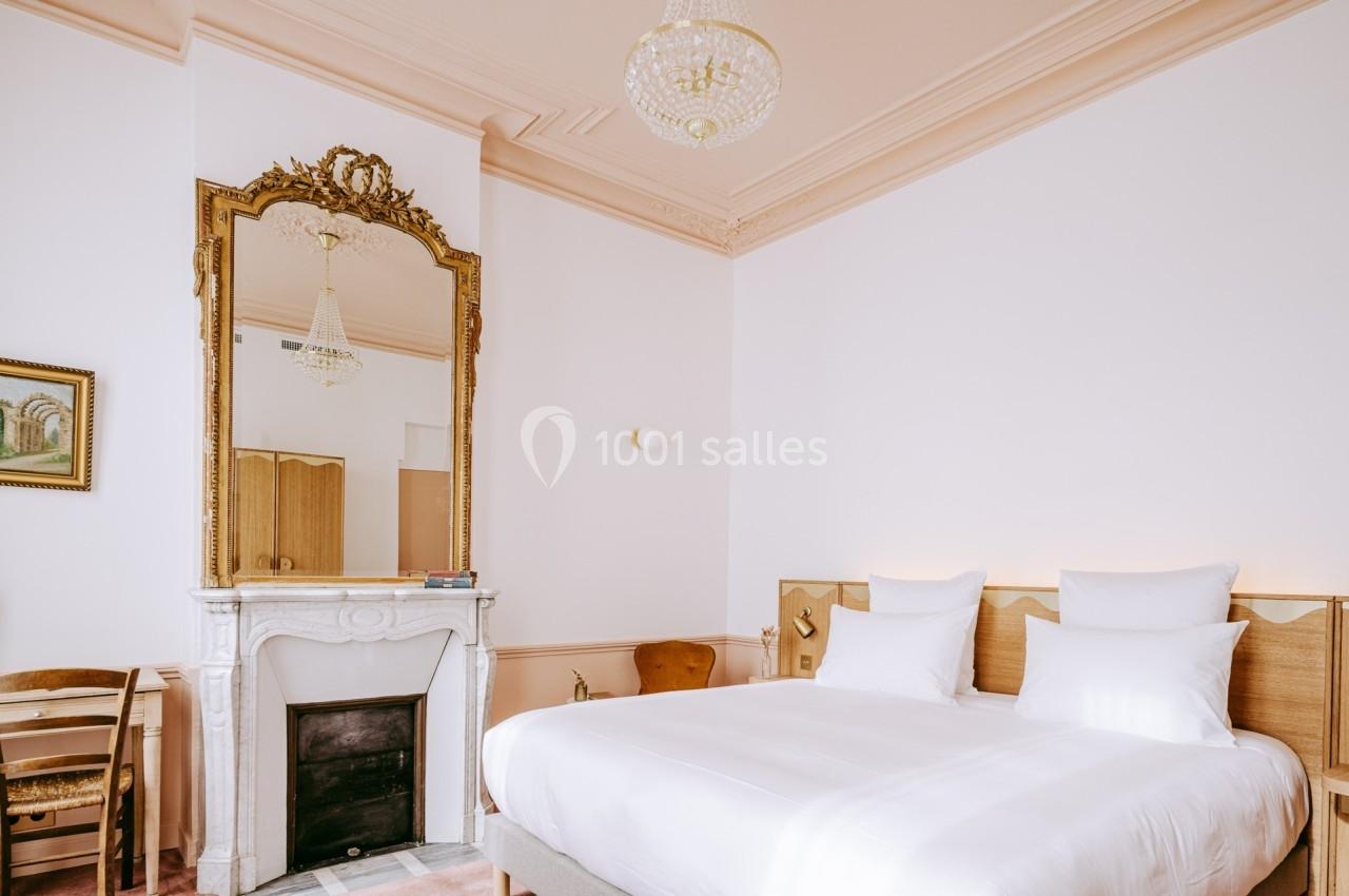 Chambre lumineuse avec lit double, cheminée en marbre, grand miroir doré et lustre suspendu.