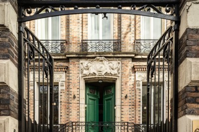 Façade d'une maison en briques avec toit mansardé, portail en fer forgé et arbres dénudés en premier plan.