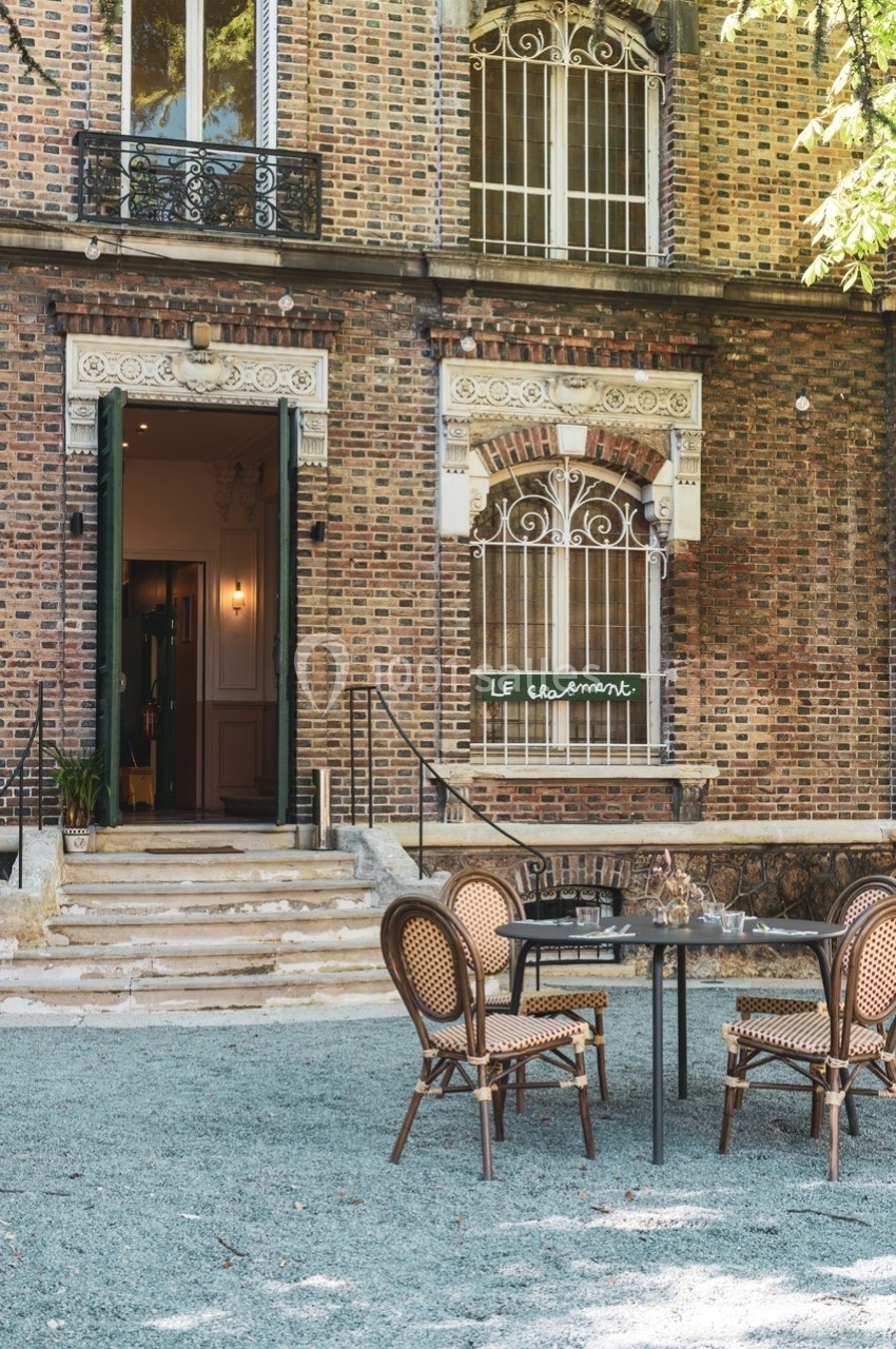 Façade en briques d'un bâtiment avec une terrasse aménagée, des tables et chaises en rotin devant l'entrée.