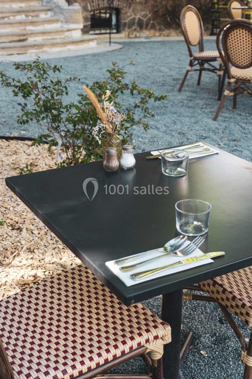 Table extérieure avec deux chaises en osier, dressée avec couverts, verres et un petit bouquet décoratif.