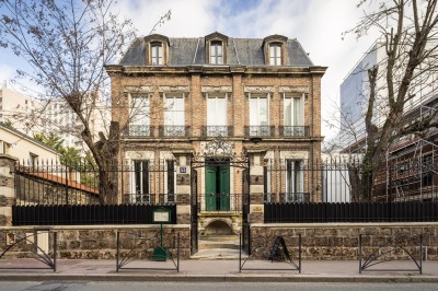 Façade d'une maison en briques avec toit mansardé, portail en fer forgé et arbres dénudés en premier plan.