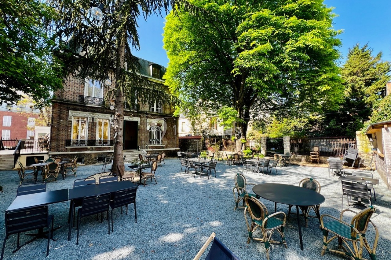 Terrasse extérieure avec tables et chaises sous des arbres, devant une maison en briques par une journée ensoleillée.