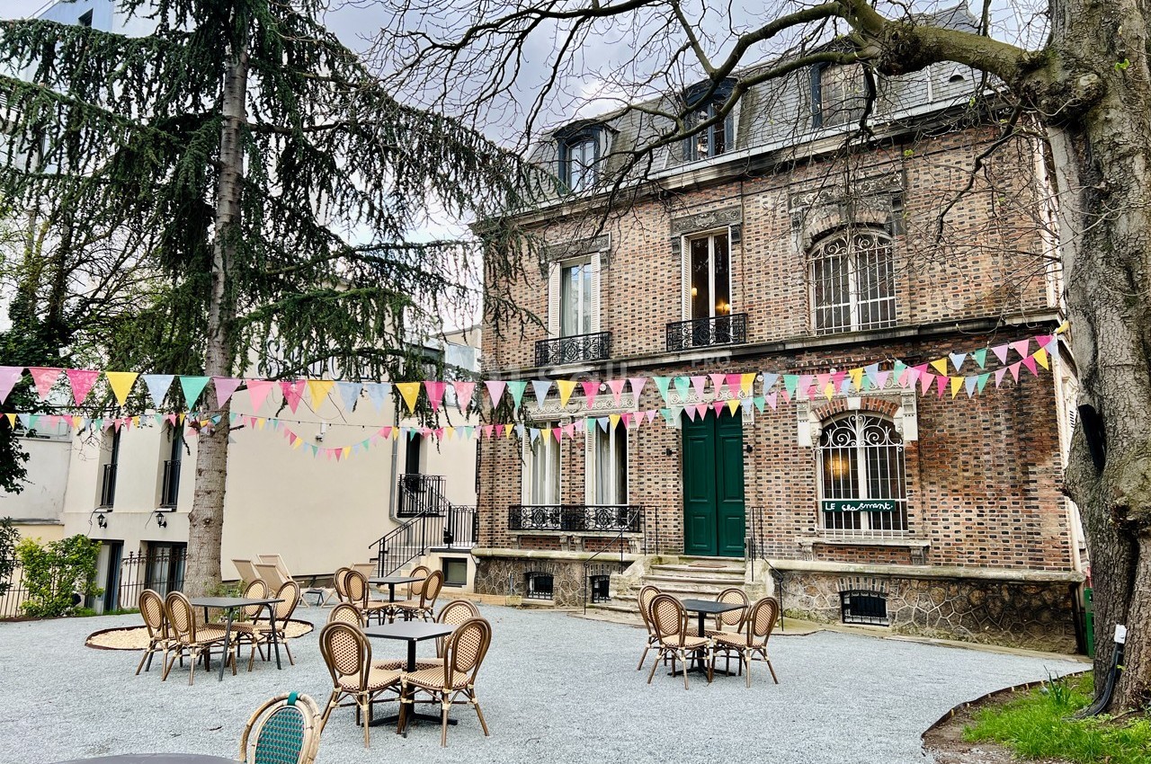 Cour extérieure avec tables et chaises en rotin, décorée de guirlandes colorées, devant une maison en briques.