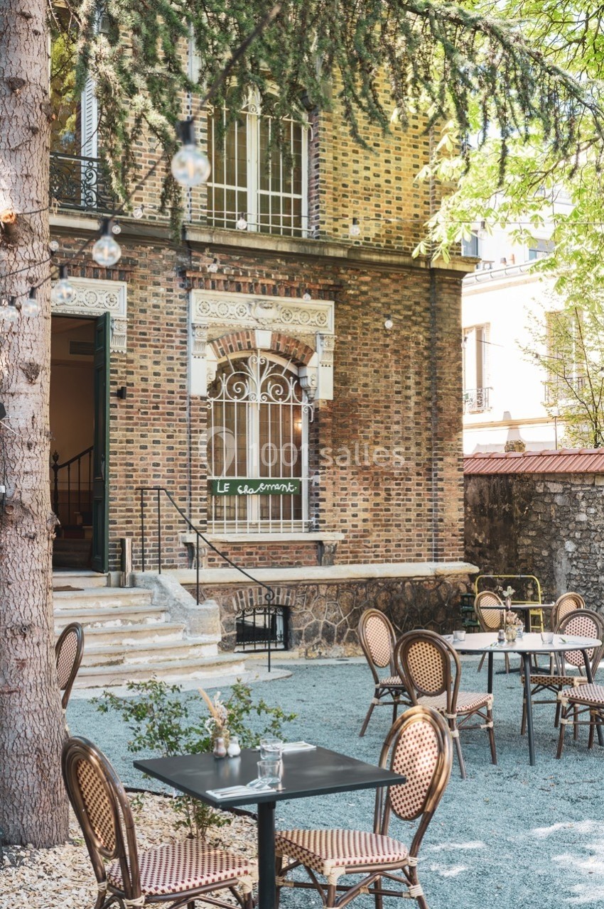 Terrasse extérieure avec tables et chaises en rotin devant une façade en briques, entourée de verdure.