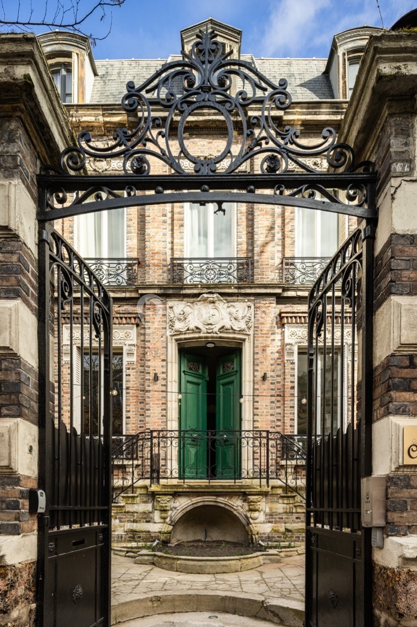 Portail en fer forgé ouvert donnant sur une façade en briques avec balcon et porte d'entrée verte.