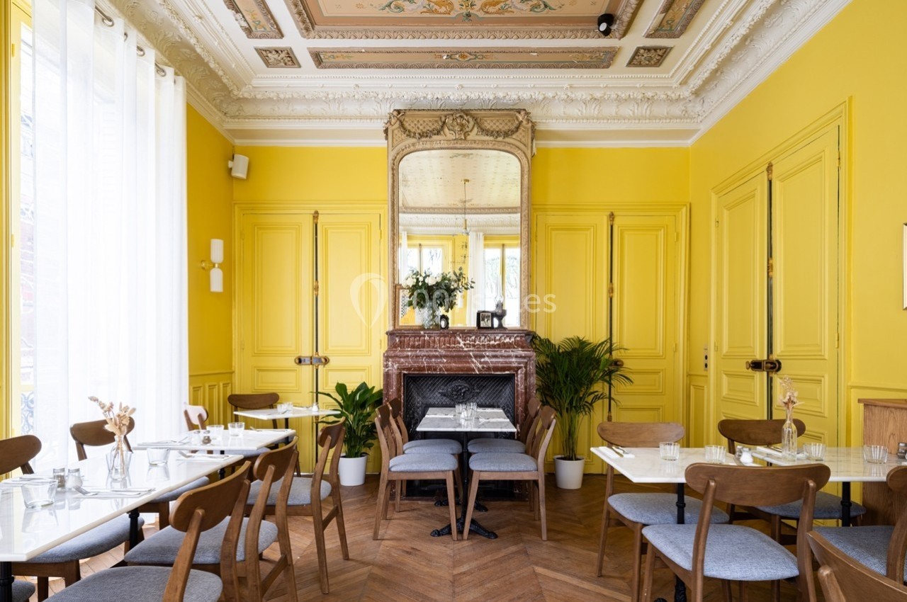 Salle de restaurant lumineuse avec murs jaunes, cheminée en marbre, miroir ancien et tables dressées.