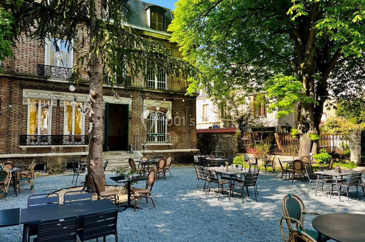Cour extérieure avec tables et chaises disposées sous des arbres, devant une maison en briques avec des fenêtres ornées.