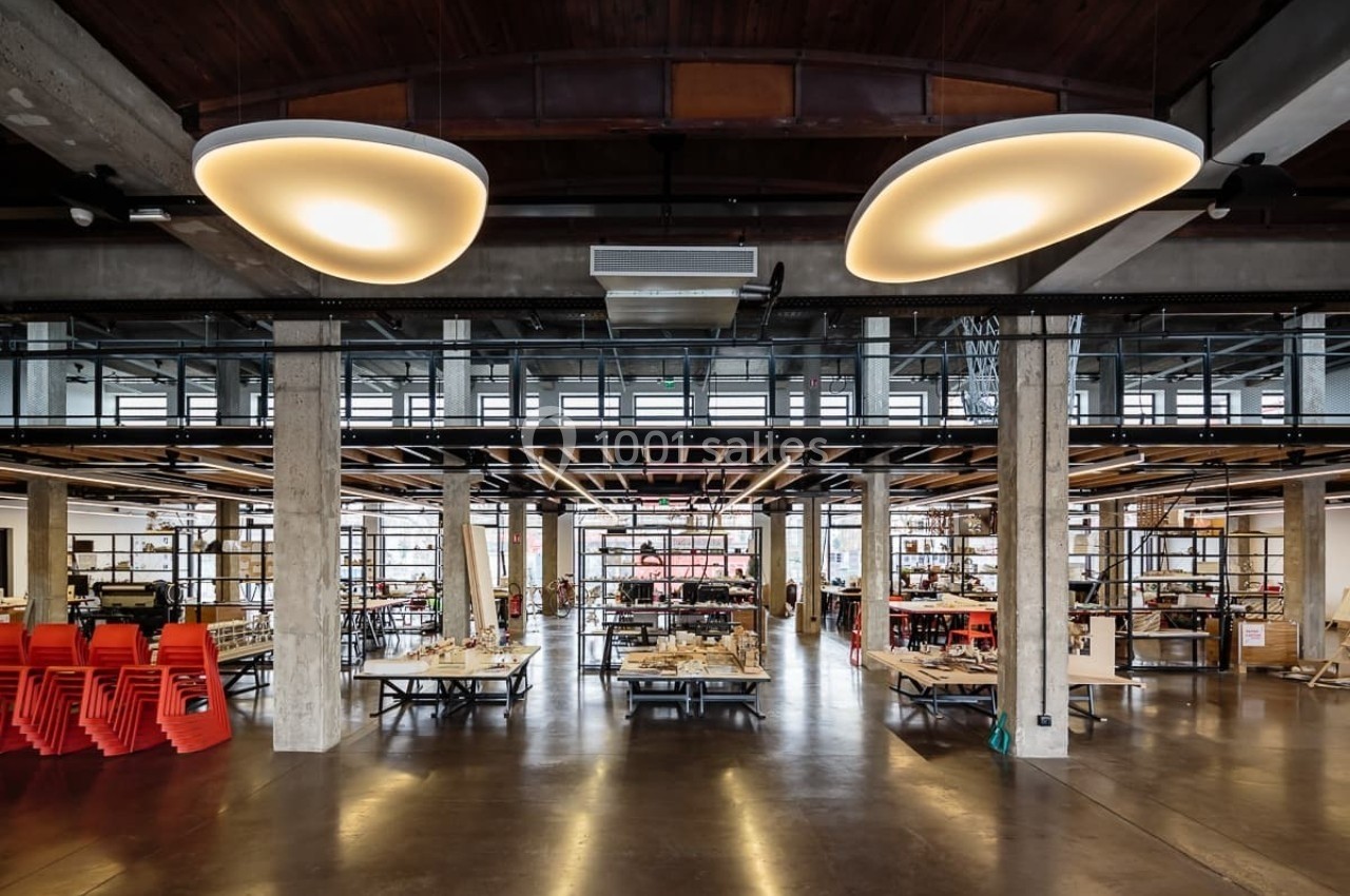 Grand espace intérieur industriel avec mezzanine, tables de travail, chaises empilées et éclairage suspendu.