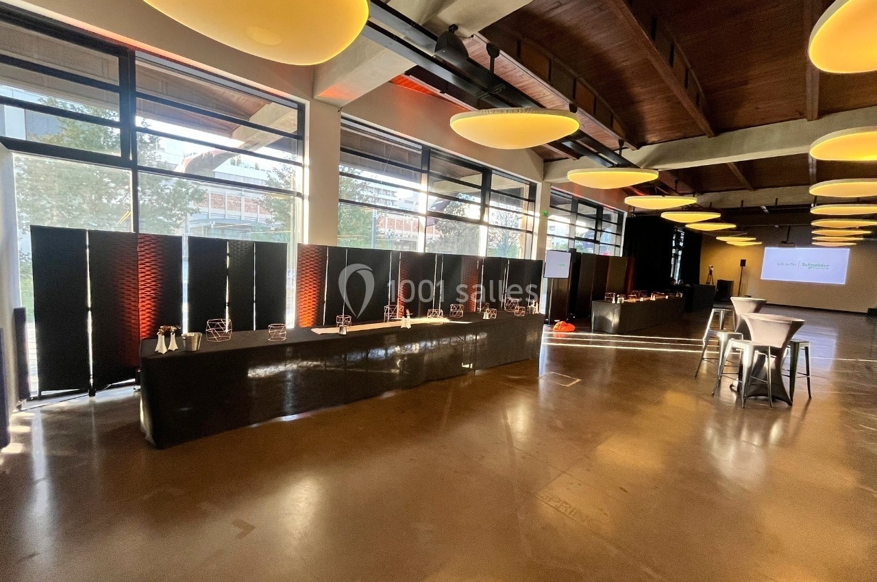 Salle lumineuse avec de grandes baies vitrées, tables hautes et buffet disposé le long d'un mur.