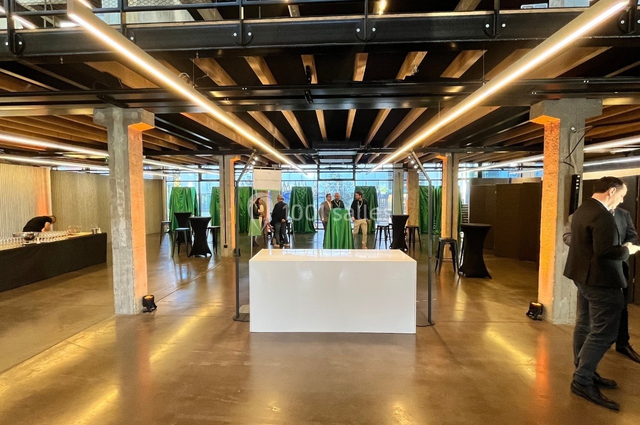 Salle moderne avec plafond en bois, tables hautes, buffet sur la gauche et quelques personnes discutant au fond.