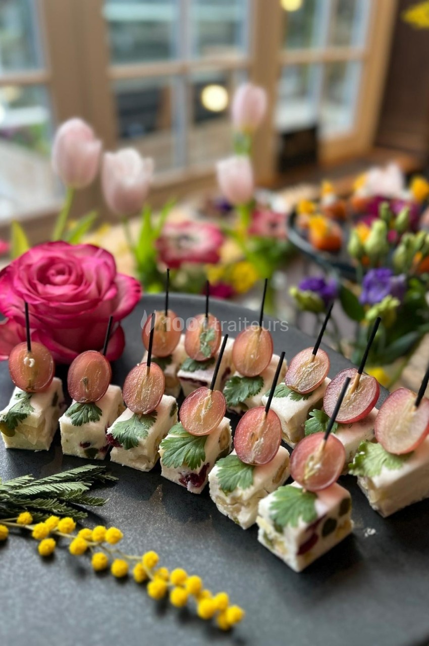 Plateau de bouchées apéritives avec fromage, raisins et herbes fraîches, décoré de fleurs en arrière-plan.