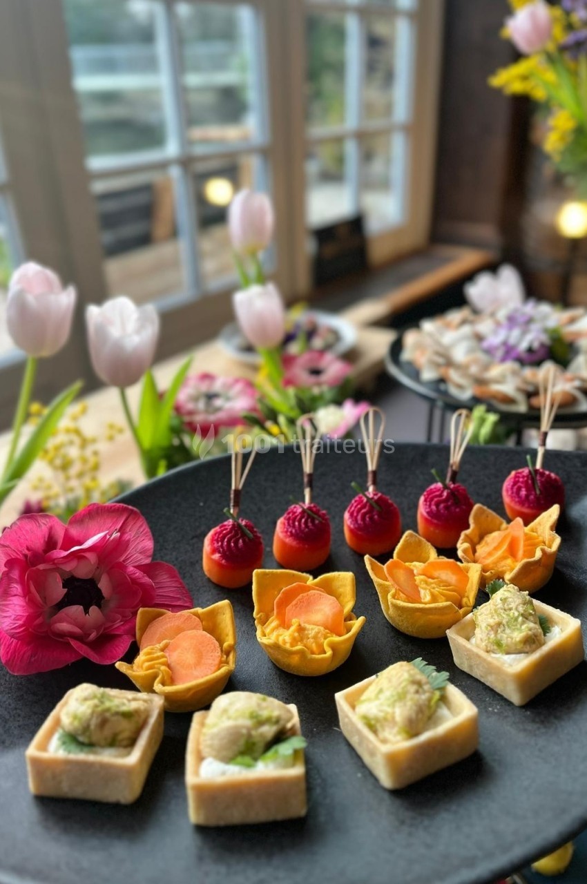 Plateau de bouchées apéritives variées, disposées sur un plat noir, avec des fleurs en arrière-plan près d'une fenêtre.