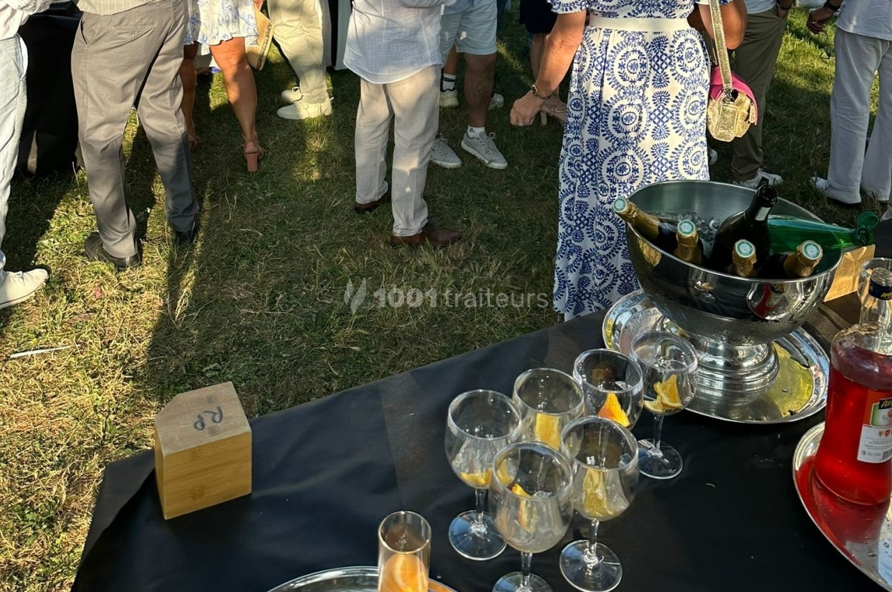 Des verres de cocktail et une seau à champagne sur une table noire lors d'un événement en plein air avec des invités debout.