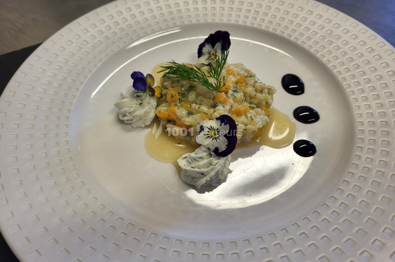 Tartare de poisson dressé avec fleurs comestibles, crème herbacée et sauce, présenté sur une assiette blanche texturée.