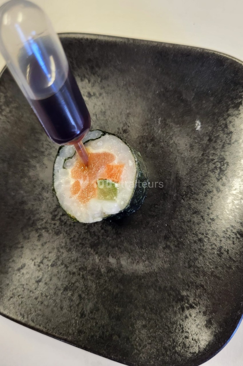 Rouleau de sushi garni de saumon et avocat, présenté avec une pipette contenant une sauce foncée.
